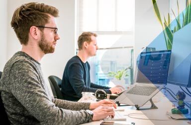 Two men at desk to illustrate article on hiring remote employees in other countries. By Tim Van Der Kuip on Unsplash.